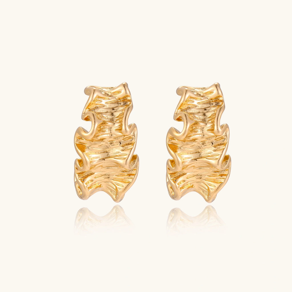Pair of gold huggie hoops with sculpted wave texture and dimensional ripples on neutral background.